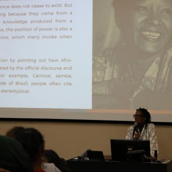 Djamila Ribeiro holds a microphone. Behind her there is an image of Lelia Gonzalez